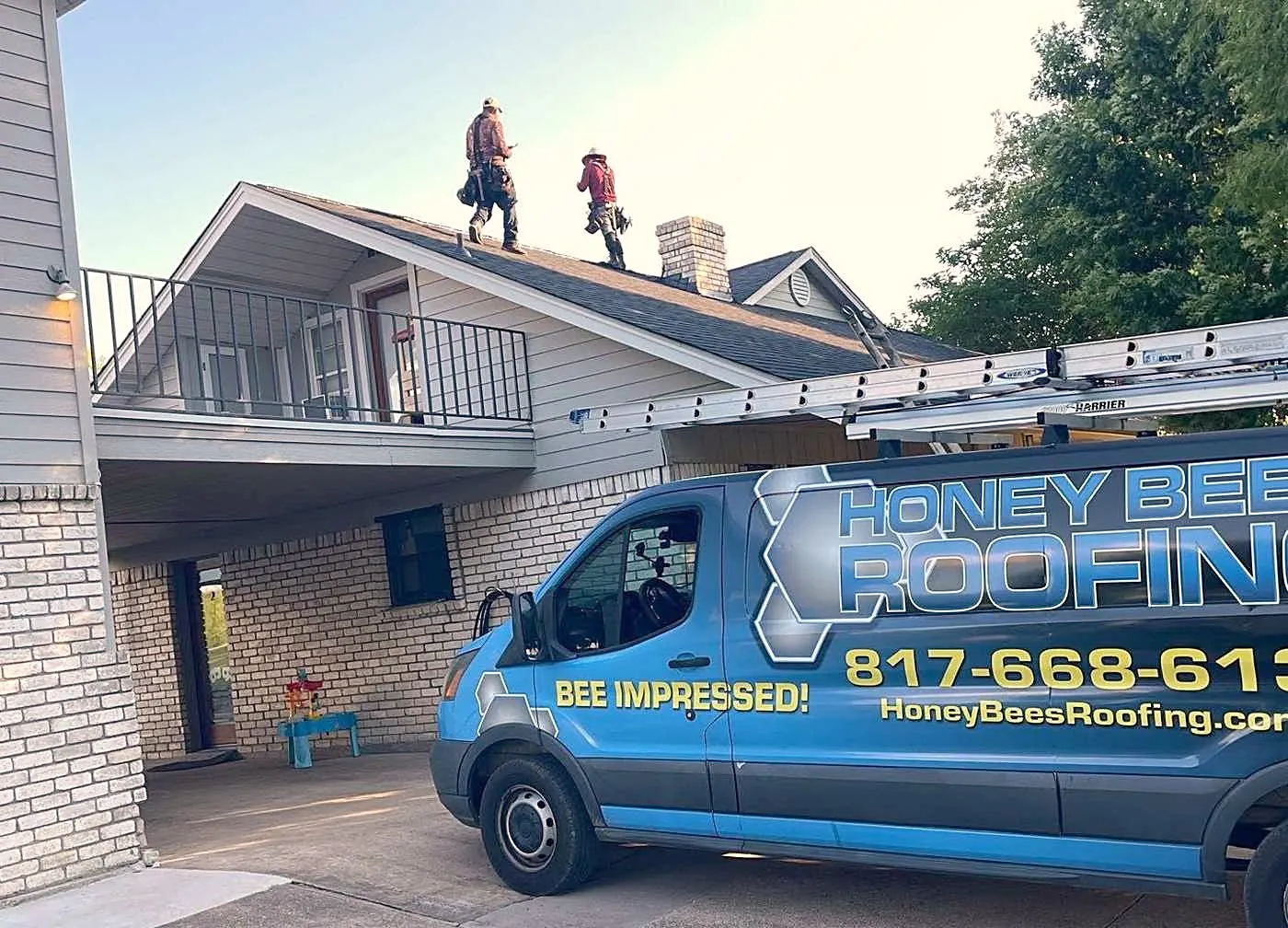 A Honey Bees Roofing van with "Bee Impressed!" on the side, parked at a home while two roofers walk the ridge