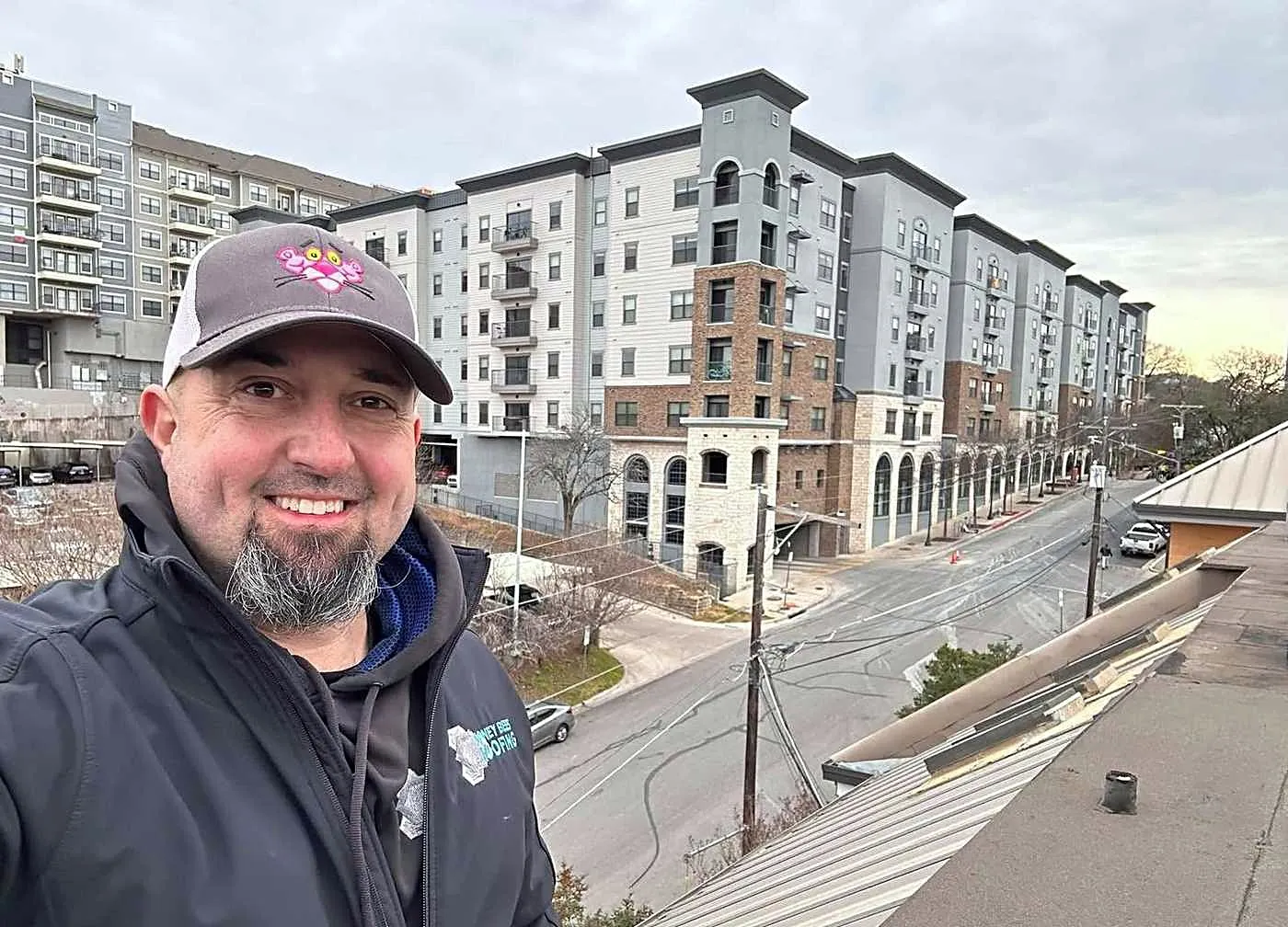 Daniel Pentikis, owner of Honey Bees Roofing, on a metal roof with apartment buildings behind him