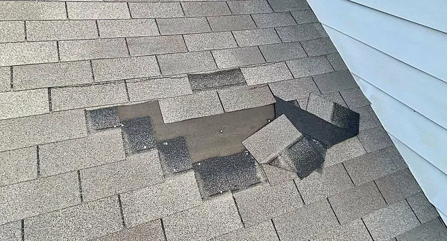 Torn and lifted shingle tabs on a roof beside a wall